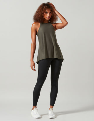 A woman modeling a sleeveless olive green athletic tank top paired with black leggings and white sneakers, standing against a light gray studio background.