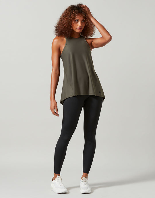 A woman modeling a sleeveless olive green athletic tank top paired with black leggings and white sneakers, standing against a light gray studio background.