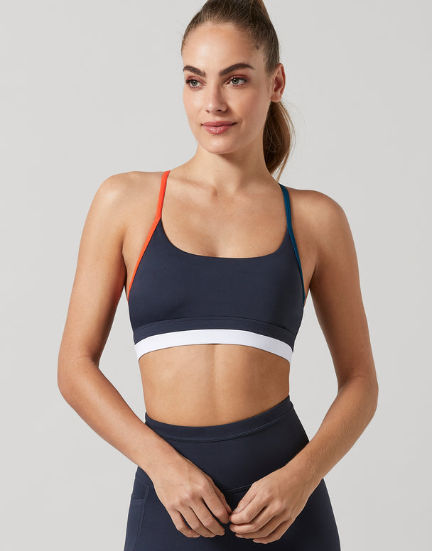 Woman wearing a navy sports bra with white band and orange straps on a light gray background