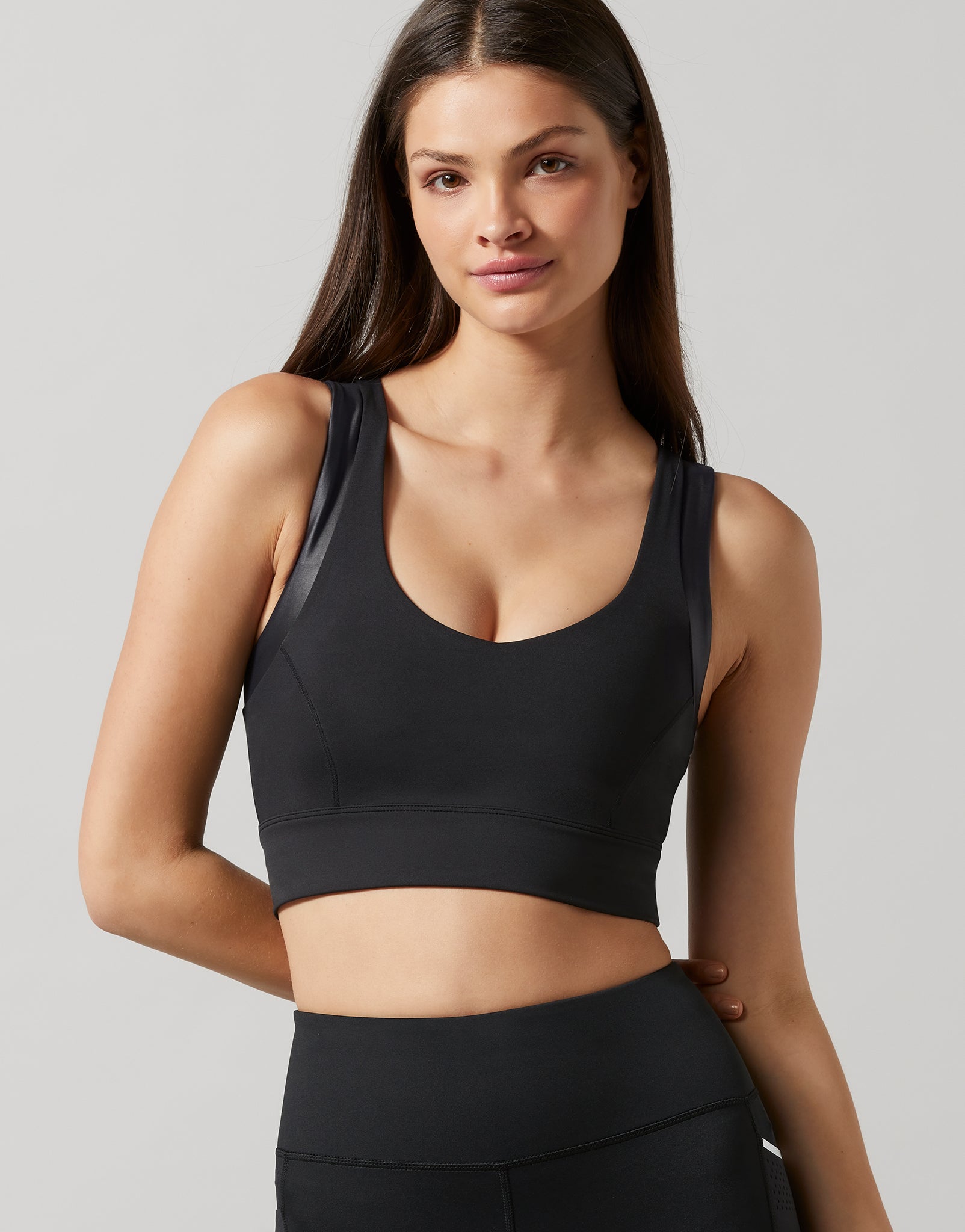 A woman modeling a black athletic sports bra with sheer mesh side panels, paired with black athletic bottoms, against a solid light gray background.