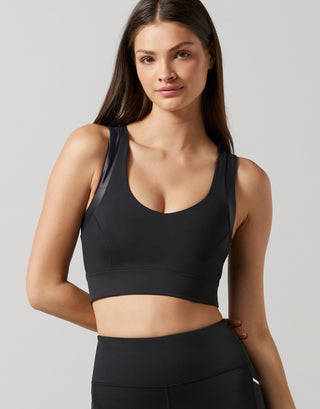 A woman modeling a black athletic sports bra with sheer mesh side panels, paired with black athletic bottoms, against a solid light gray background.