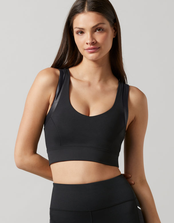 A woman modeling a black athletic sports bra with sheer mesh side panels, paired with black athletic bottoms, against a solid light gray background.