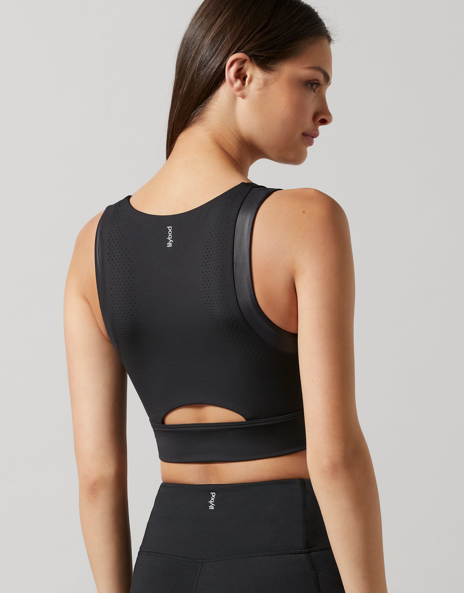 A woman shown from the back wearing a black athletic sports bra with a small white logo and a cutout at the waist, paired with matching black bottoms, against a light gray studio background.
