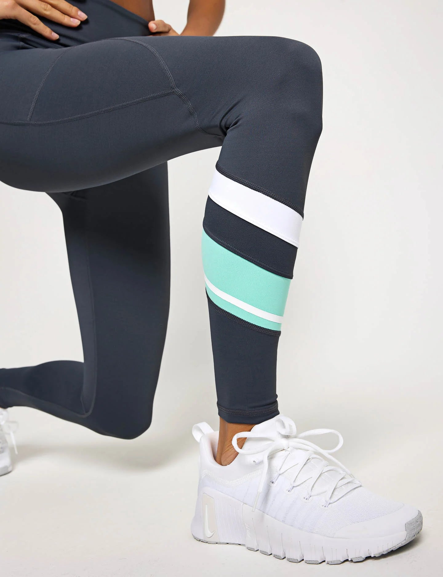 Person wearing dark gray leggings with white and light blue stripes, paired with white sneakers on a light background.