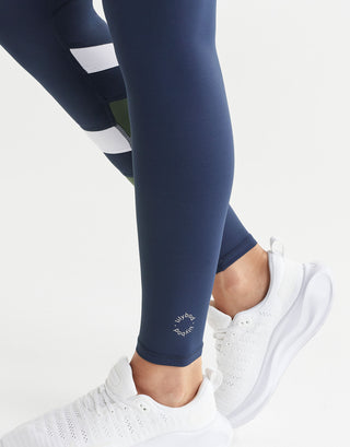 Close up of navy leggings with white stripes worn by a person, paired with white sneakers on a light grey background.
