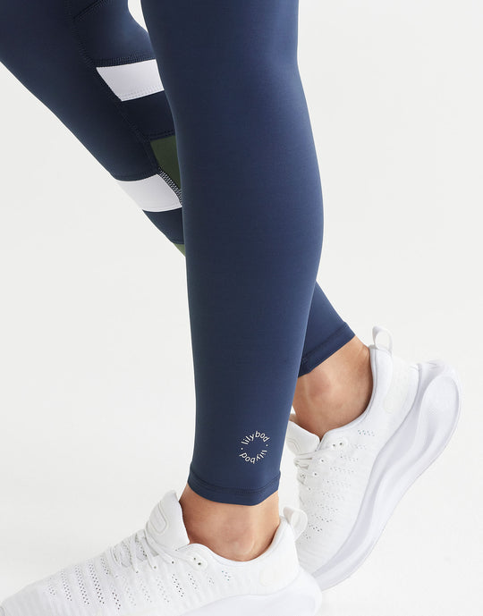 Close up of navy leggings with white stripes worn by a person, paired with white sneakers on a light grey background.
