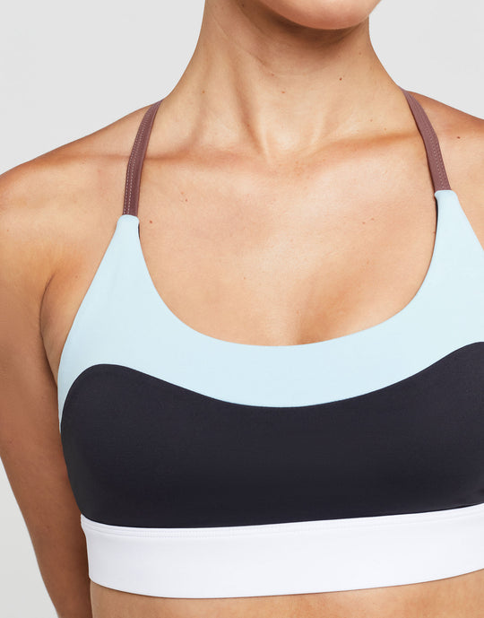 A model wearing a sports bra with color blocked accent stripes in charcoal and pale blue with a white scoop neck.