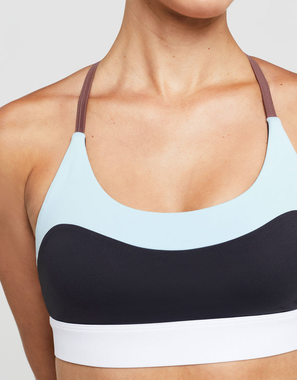 A model wearing a sports bra with color blocked accent stripes in charcoal and pale blue with a white scoop neck.
