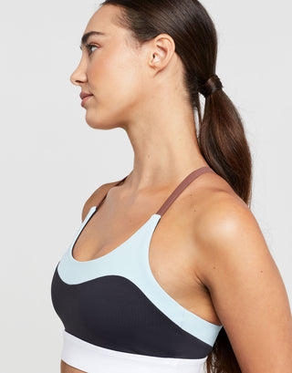 A woman wearing a sports bra with color blocked accent stripes in charcoal and pale blue with a white scoop neck.