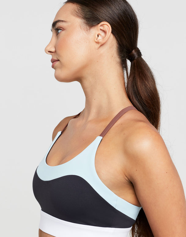 A woman wearing a sports bra with color blocked accent stripes in charcoal and pale blue with a white scoop neck.