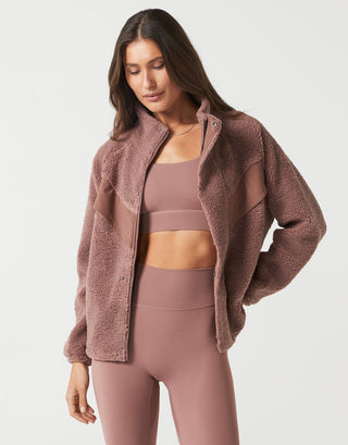 A woman modeling a dusty rose fleece zip-up jacket over a matching dusty rose sports bra and leggings set, photographed against a clean light gray studio background.