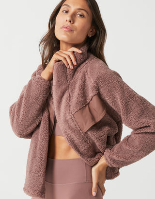 A woman wearing a dusty mauve-brown sherpa fleece cropped jacket with a high collar and a matching sports bra and leggings set, photographed against a clean light gray studio background.