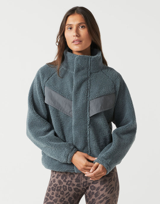 A woman wearing a textured fleece jacket in a muted slate blue-gray color with contrasting dark gray panels on the chest, paired with leopard-print leggings, against a plain light gray studio background.