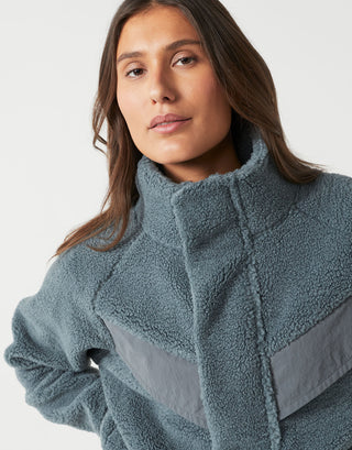 A close-up of a woman wearing a dusty slate blue sherpa fleece jacket with a high collar and contrasting smooth gray paneling on the front.