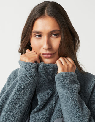 A close-up of a woman wearing a textured, plush fleece pullover sweater in a muted slate blue-gray color, posing with her hands near her collar against a light gray background.
