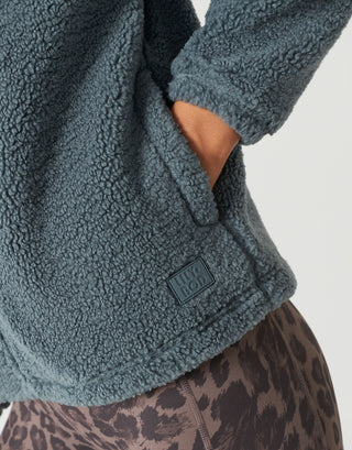 Close-up of a person wearing a textured fleece jacket in a muted blue-gray color, with a small rectangular brand patch reading UGG LOFT visible near the hem. The photo is cropped to show the jacket fabric, sleeve cuff, hand in pocket, and part of leopard-print bottoms against a light neutral background.