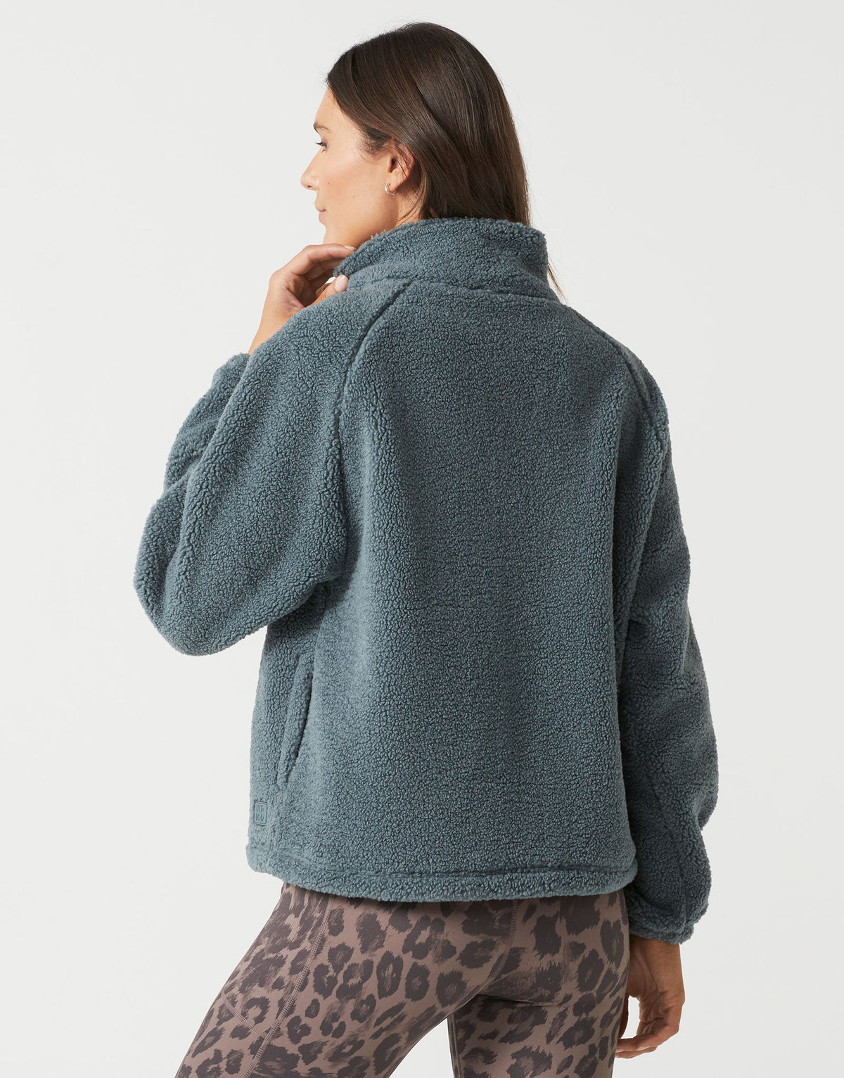 A woman shown from the back wearing a dusty slate blue sherpa fleece jacket and leopard print leggings against a light gray studio background.