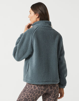 A woman shown from the back wearing a dusty slate blue sherpa fleece jacket and leopard print leggings against a light gray studio background.