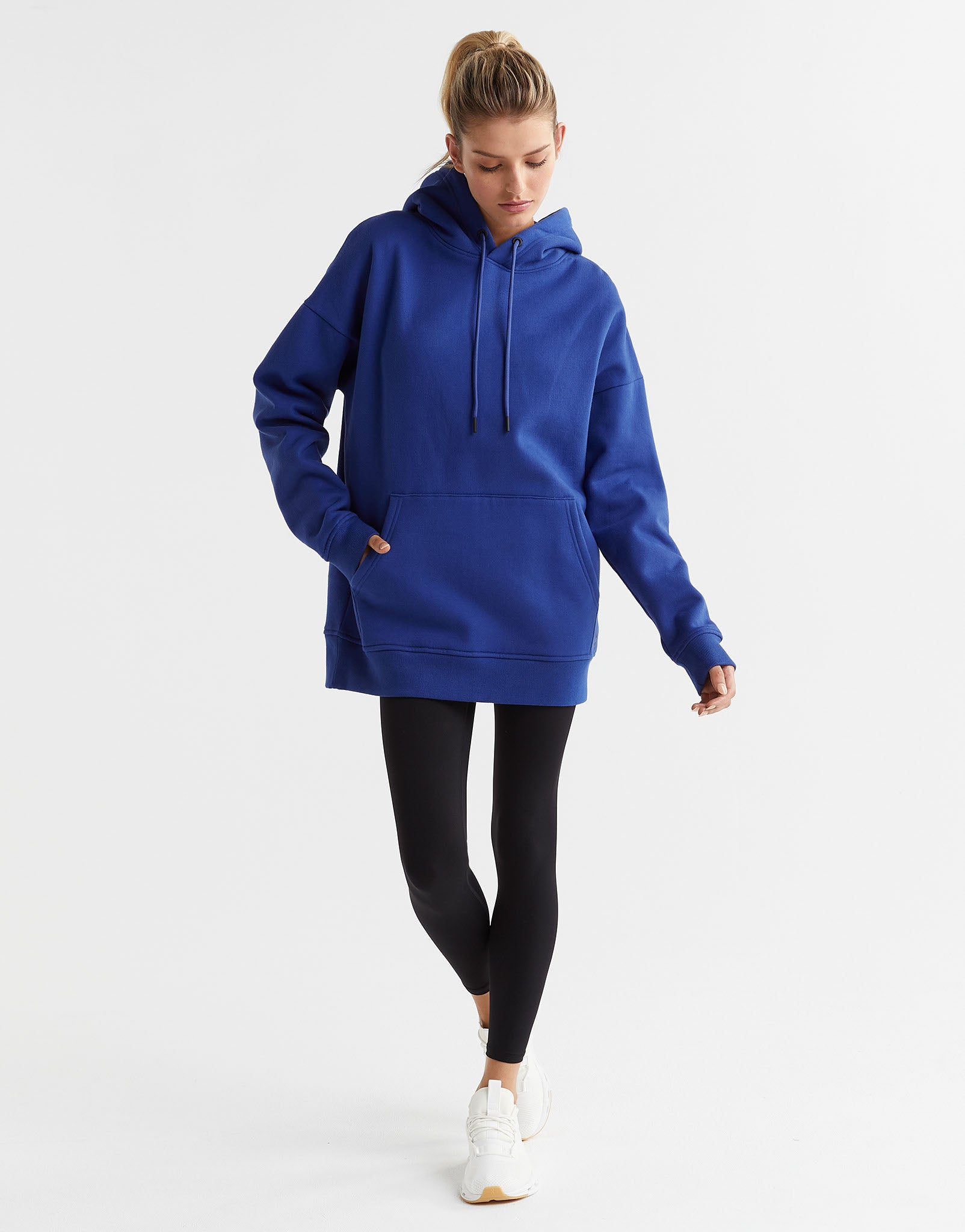 A woman wearing a blue hooded fleece sweater with a kangaroo pocket and drawstrings on the hood. The sweater is oversized and she is also wearing black leggings and white shoes.
