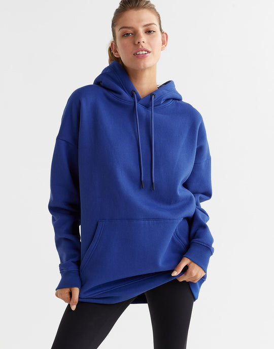 A woman wearing a blue hooded fleece sweater with a kangaroo pocket and drawstrings on the hood. The sweater is oversized and she is also wearing black leggings.