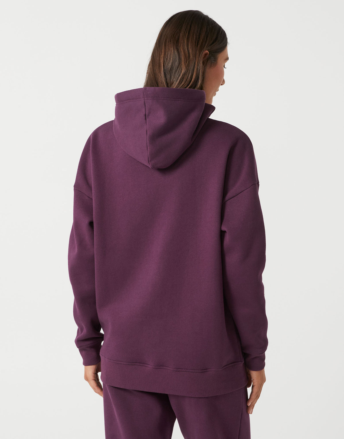 A woman viewed from the back wearing an oversized plum purple hoodie and matching sweatpants against a light gray studio background.