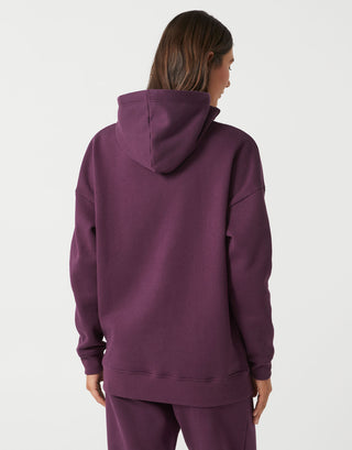 A woman viewed from the back wearing an oversized plum purple hoodie and matching sweatpants against a light gray studio background.