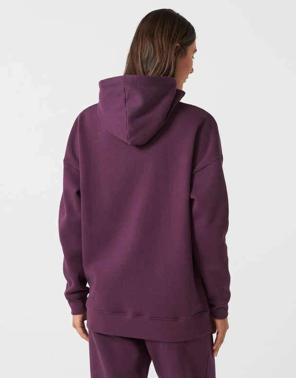 A woman viewed from the back wearing an oversized plum purple hoodie and matching sweatpants against a light gray studio background.