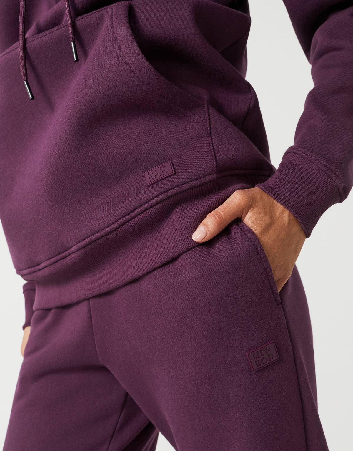 Close-up of a person wearing a matching deep plum purple sweatshirt and jogger pants set, with a hand in the pocket of the joggers and small rectangular brand patches on both garments.