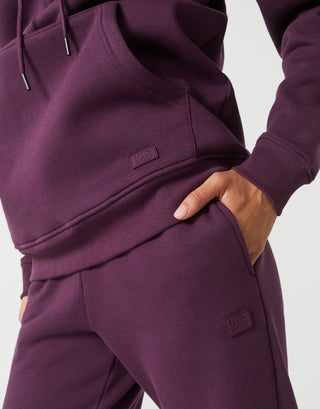 Close-up of a person wearing a matching deep plum purple sweatshirt and jogger pants set, with a hand in the pocket of the joggers and small rectangular brand patches on both garments.