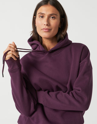 A woman wearing an oversized, deep purple pullover hoodie with matching drawstrings, shown against a plain light gray studio background.