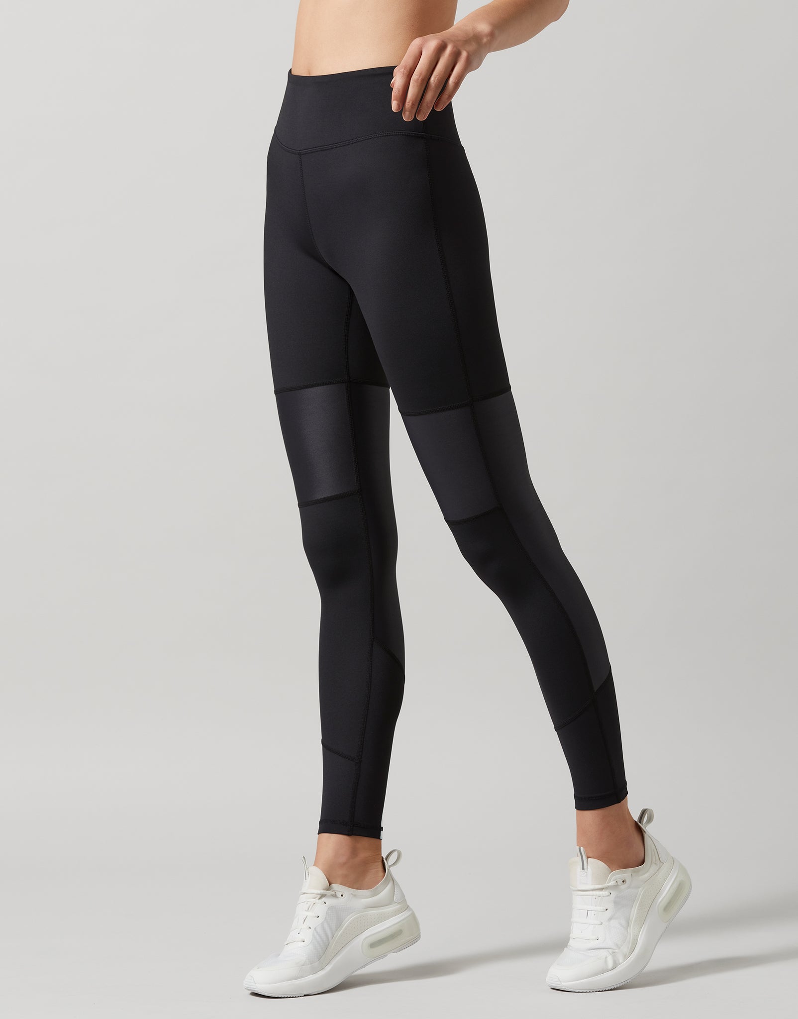 A person standing and facing to the side wearing black high-waisted leggings with a full-length design. The person is also wearing a sports bra and white sneakers.