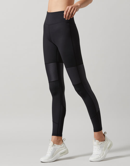 A person standing and facing to the side wearing black high-waisted leggings with a full-length design. The person is also wearing a sports bra and white sneakers.