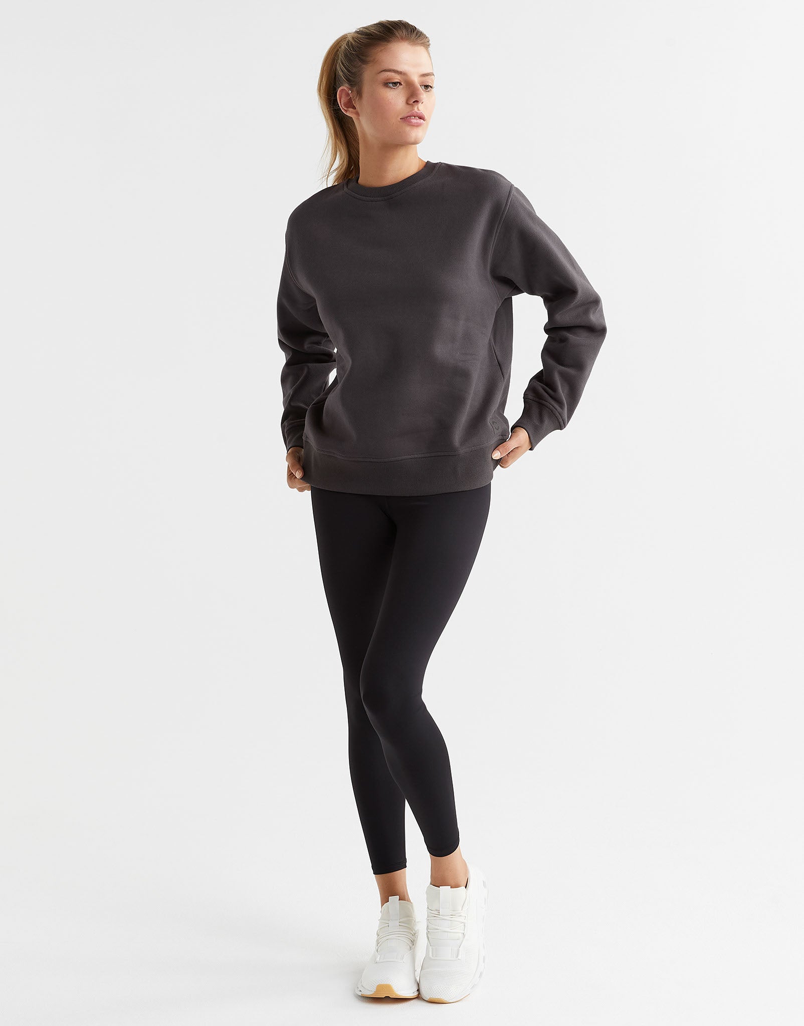 A female model wearing a gray Lilybod Montana fleece crew sweater, which is designed with a crew neck and long sleeves with black leggings and runners.