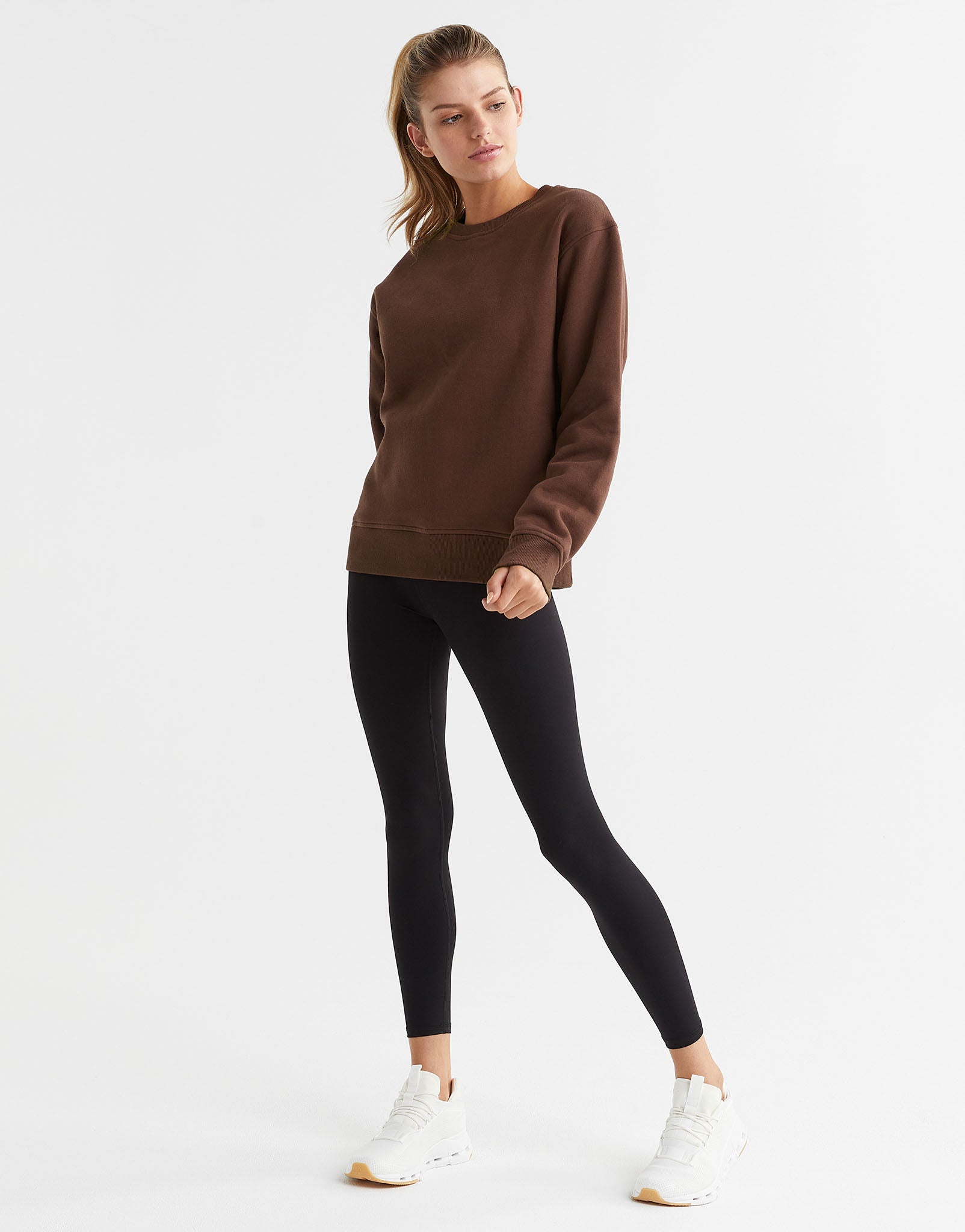 A woman wearing a brown crew neck fleece crew sweater with elasticised cuffs and a Lilybod accent logo on the front and black leggings.