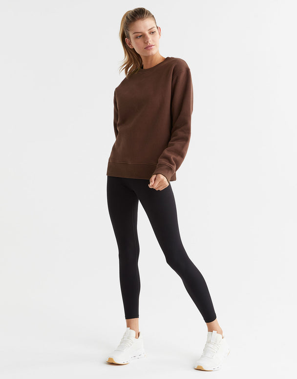 A woman wearing a brown crew neck fleece crew sweater with elasticised cuffs and a Lilybod accent logo on the front and black leggings.