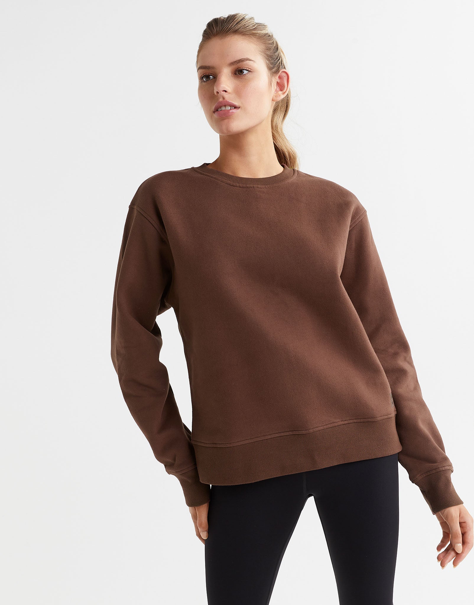 A model wearing a brown crew neck fleece crew sweater with elasticised cuffs and a Lilybod accent logo on the front.