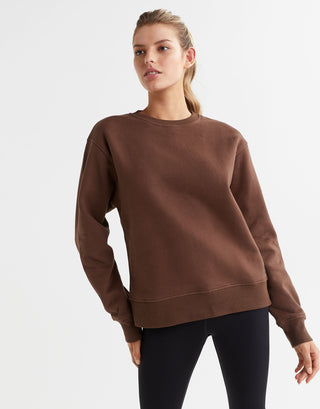 A model wearing a brown crew neck fleece crew sweater with elasticised cuffs and a Lilybod accent logo on the front.