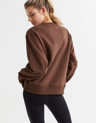 A woman wearing a brown crew neck fleece crew sweater with elasticised cuffs and a Lilybod accent logo on the front.