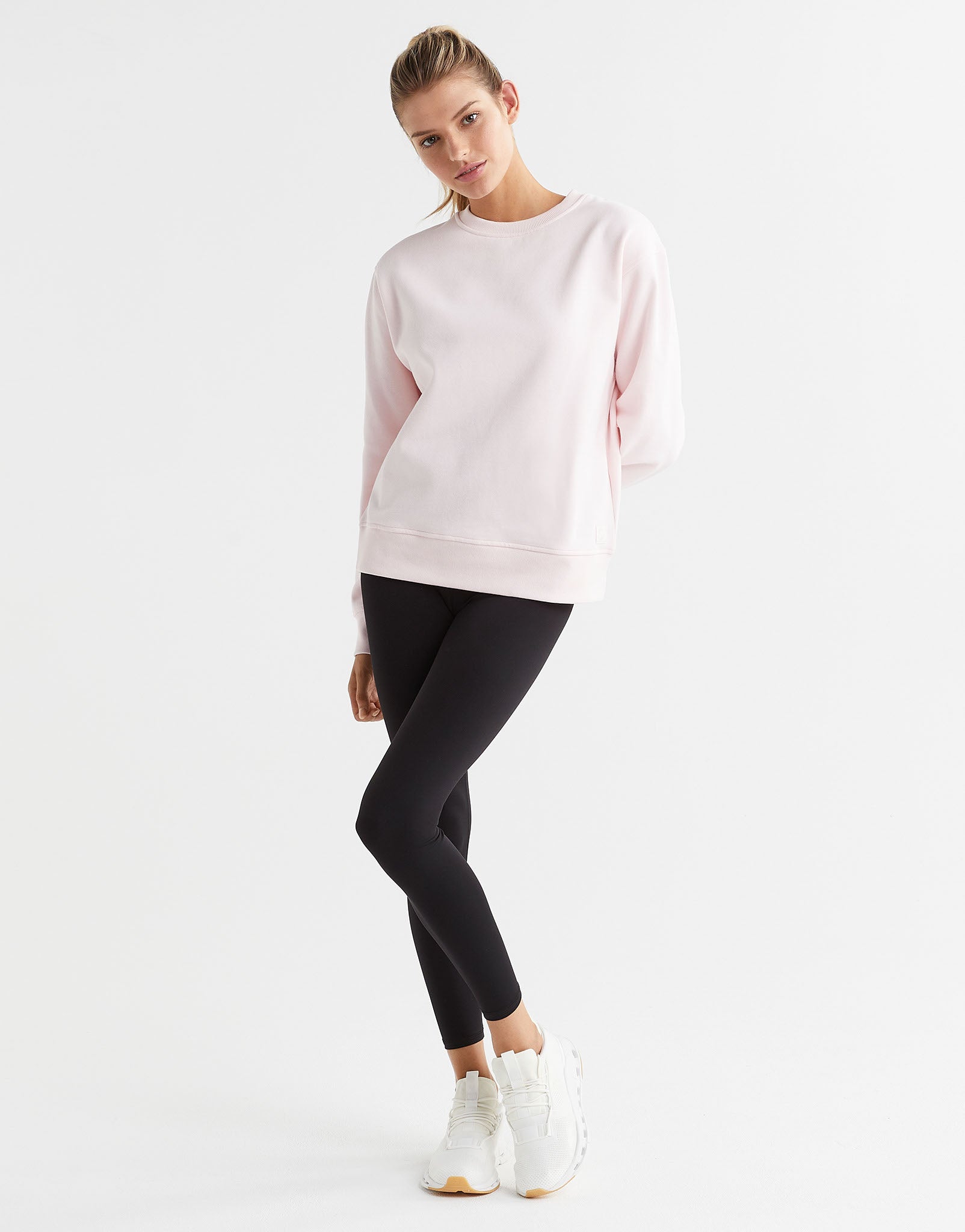 A woman wearing a light pink crew neck sweater and black leggings with white runners.