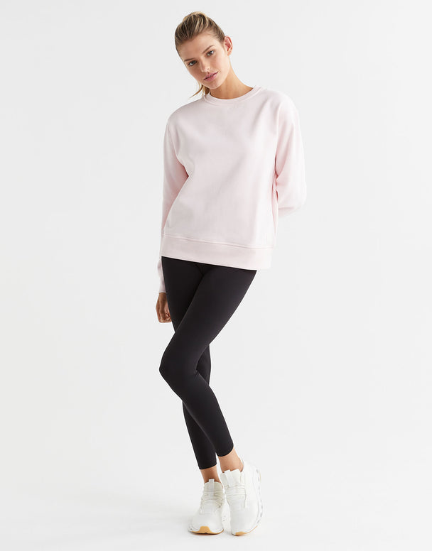 A woman wearing a light pink crew neck sweater and black leggings with white runners.