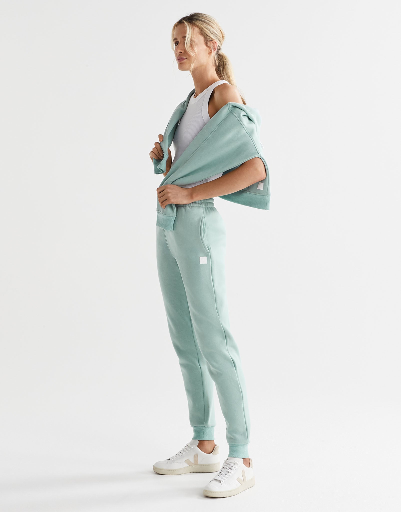 A model wearing a pair of sage green fleece-lined slim leg sweatpants with a drawstring waistband, pockets, and lilybod accent details.