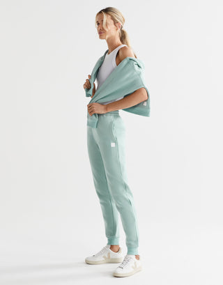 A model wearing a pair of sage green fleece-lined slim leg sweatpants with a drawstring waistband, pockets, and lilybod accent details.