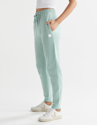 A woman in a pair of sage green fleece-lined slim leg sweatpants with a drawstring waistband, pockets, and lilybod accent details.