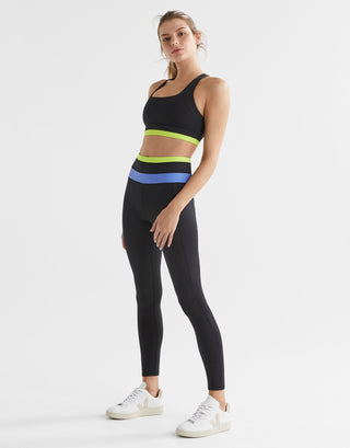A woman wearing a black sports bra with blue and yellow accents on the under-bust band and cross-back design with matching leggings.