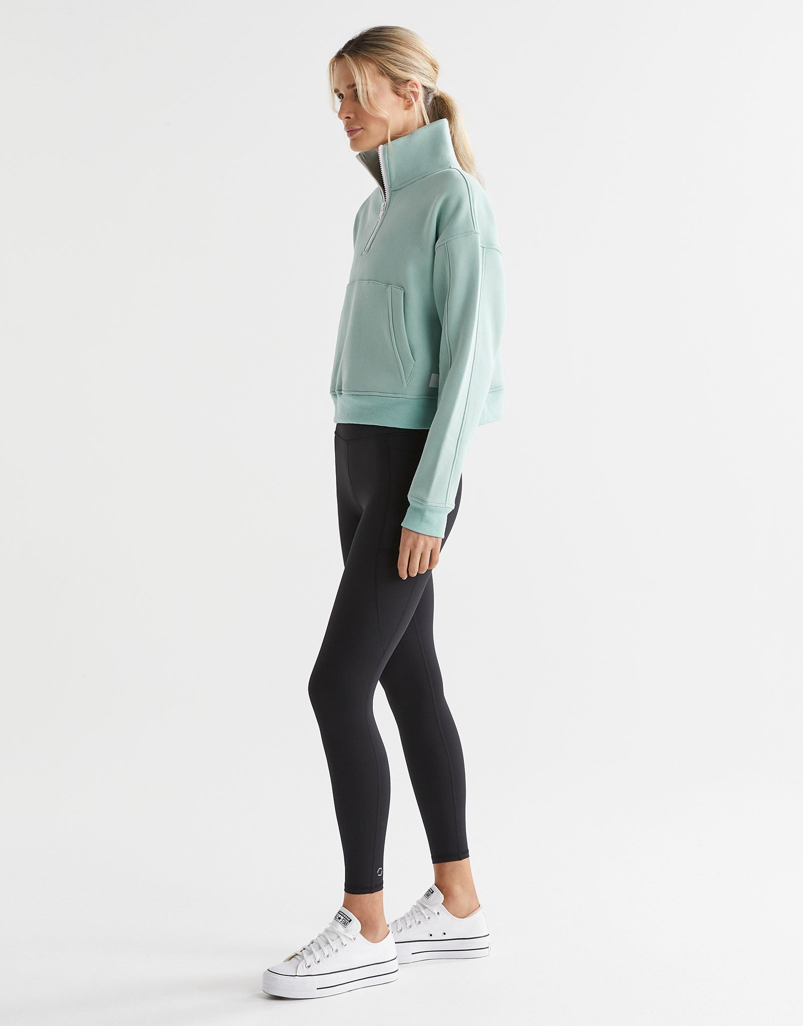 A woman wearing a sage green zip up sweater and black leggings and white runners.