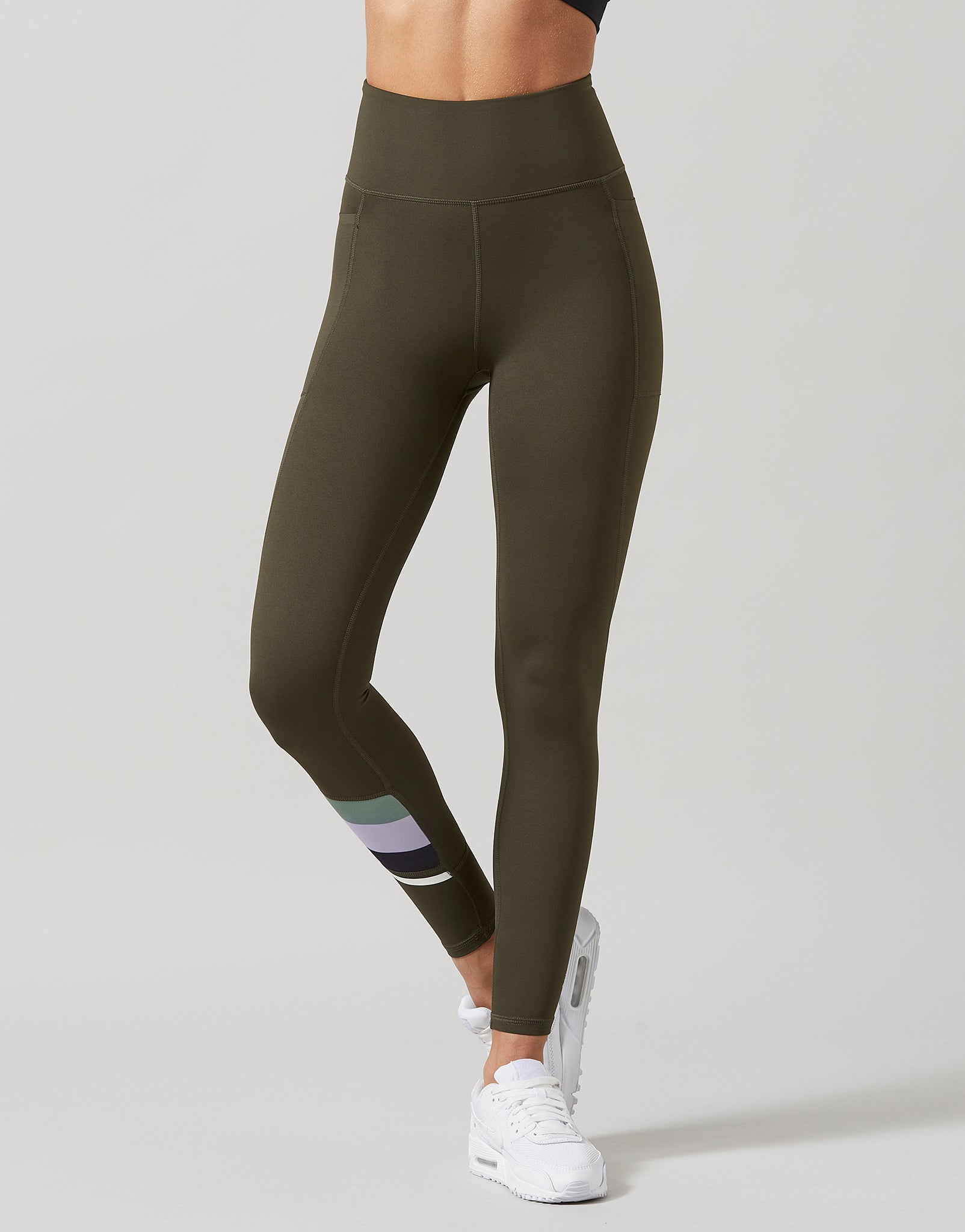 A model wearing high-waisted olive green leggings with a striped ankle accent in purple, gray, and white, paired with white sneakers, against a light gray studio background.