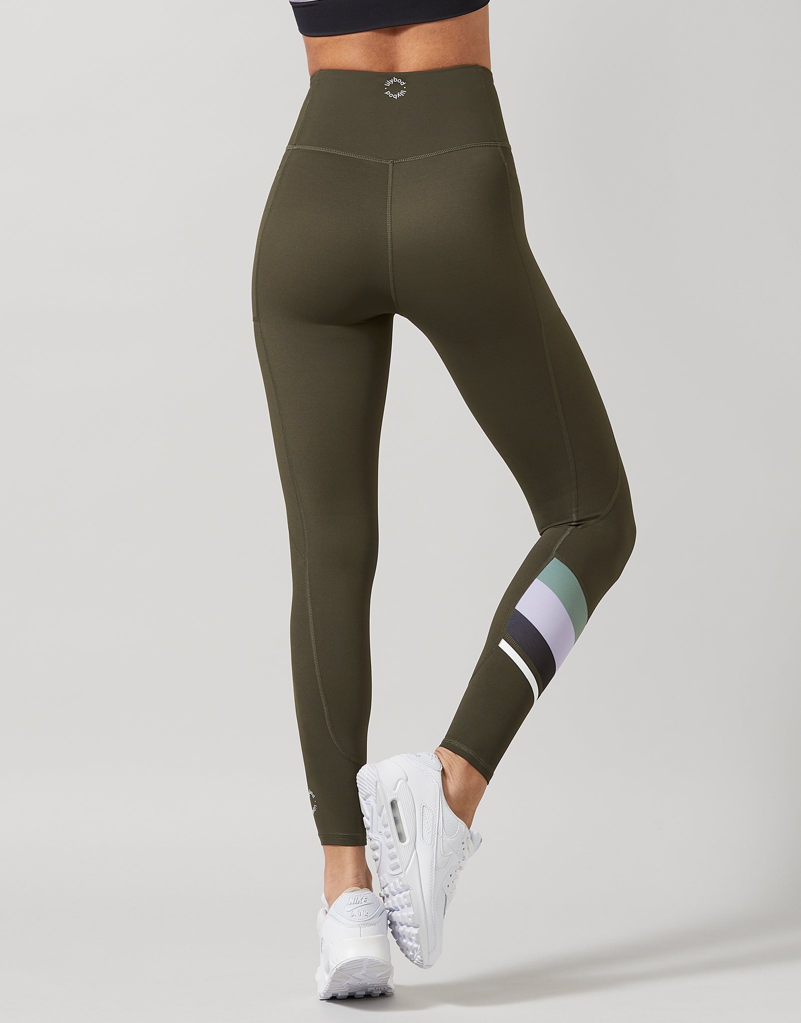 A model from the waist down wearing olive green high-waisted leggings with a geometric logo on the waistband and a striped pattern on the lower calf, paired with white sneakers against a light gray background.
