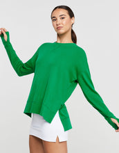 Woman wearing a green sweater and white skirt with arms outstretched on a light gray background