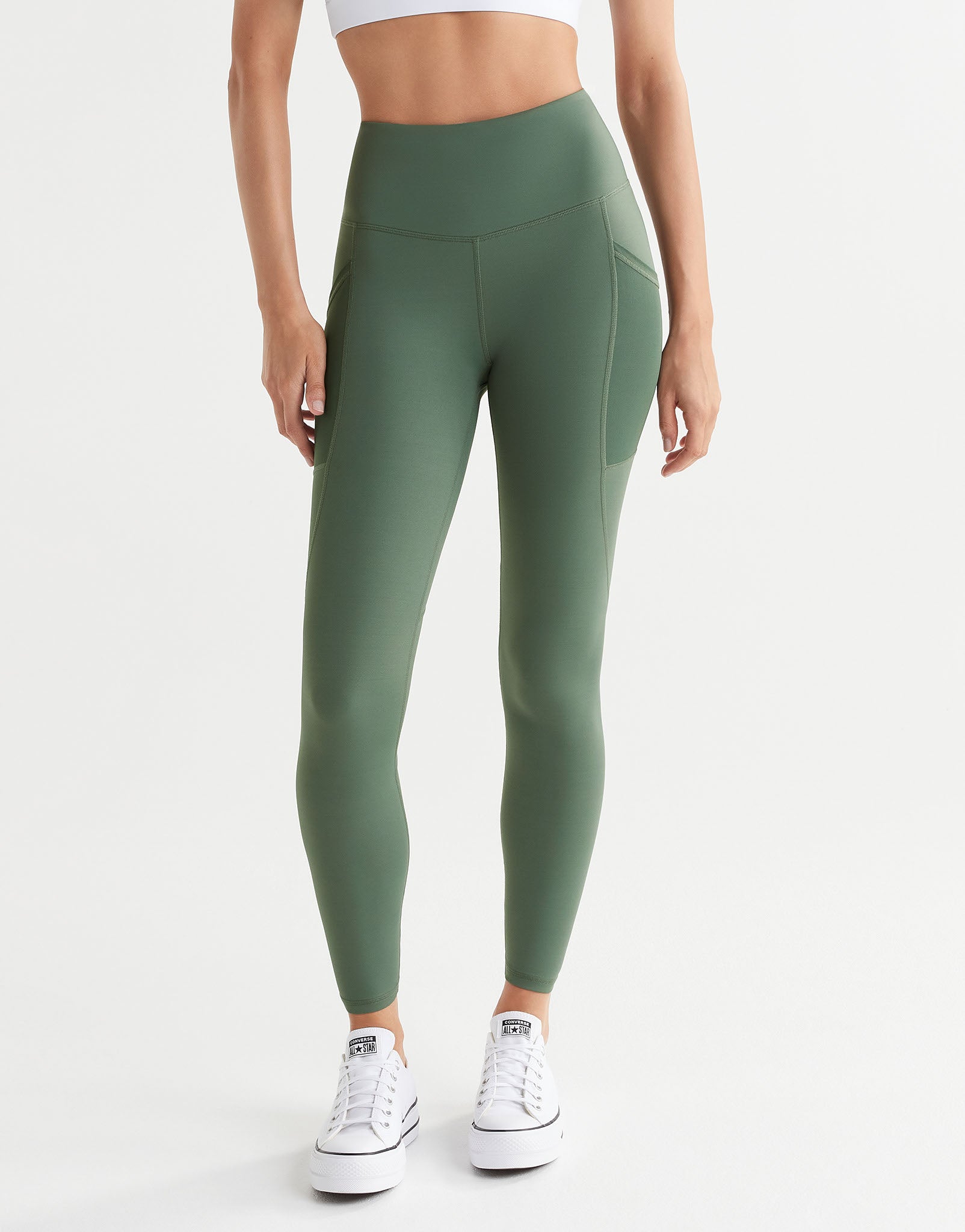 A model wearing full-length army green leggings with decorative seaming and pockets, paired with a white top and white sneakers.
