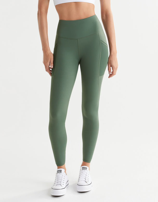 A model wearing full-length army green leggings with decorative seaming and pockets, paired with a white top and white sneakers.